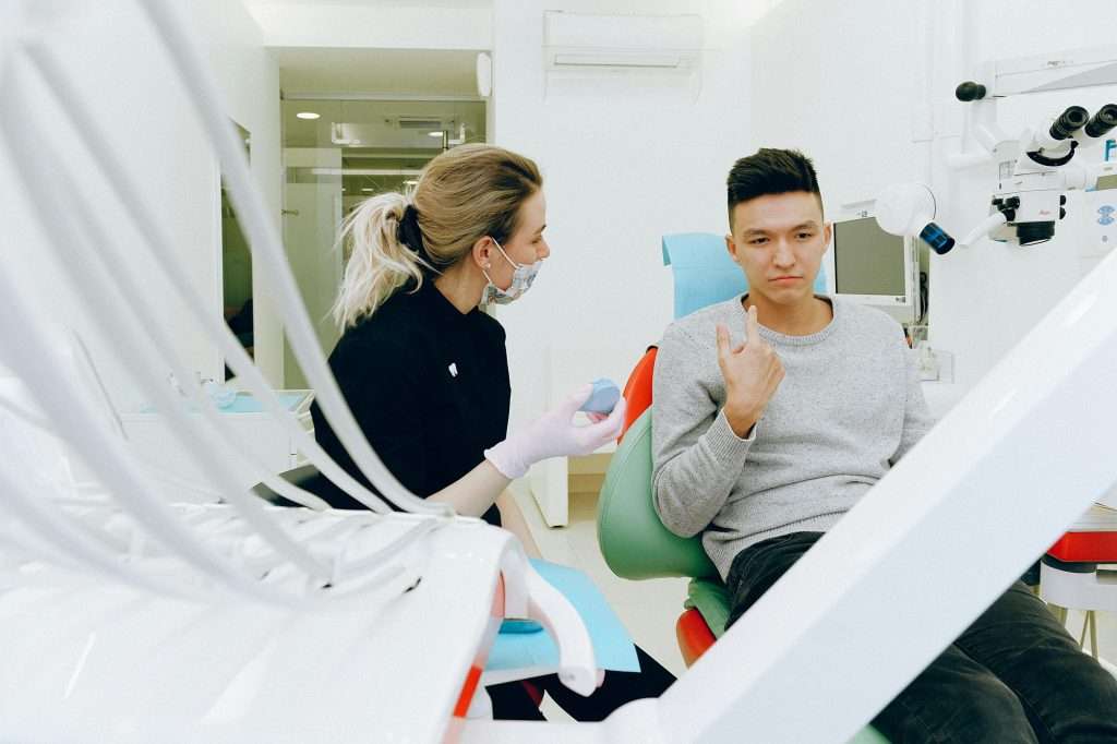 Young man pointing at his jaw as dental assistant sits nearby talking to him.